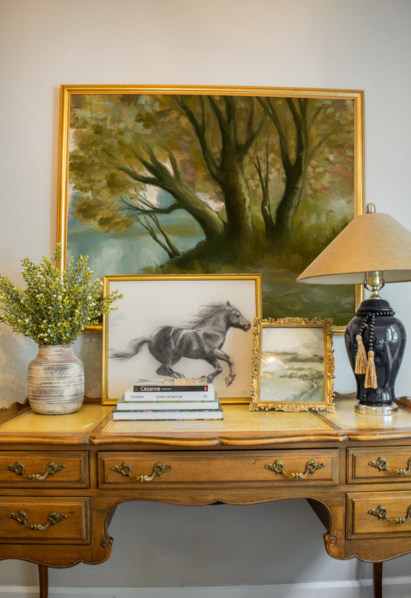 Wooden desk with decorative items including a painting of a horse, lamp, and books against a wall with a large framed landscape painting.