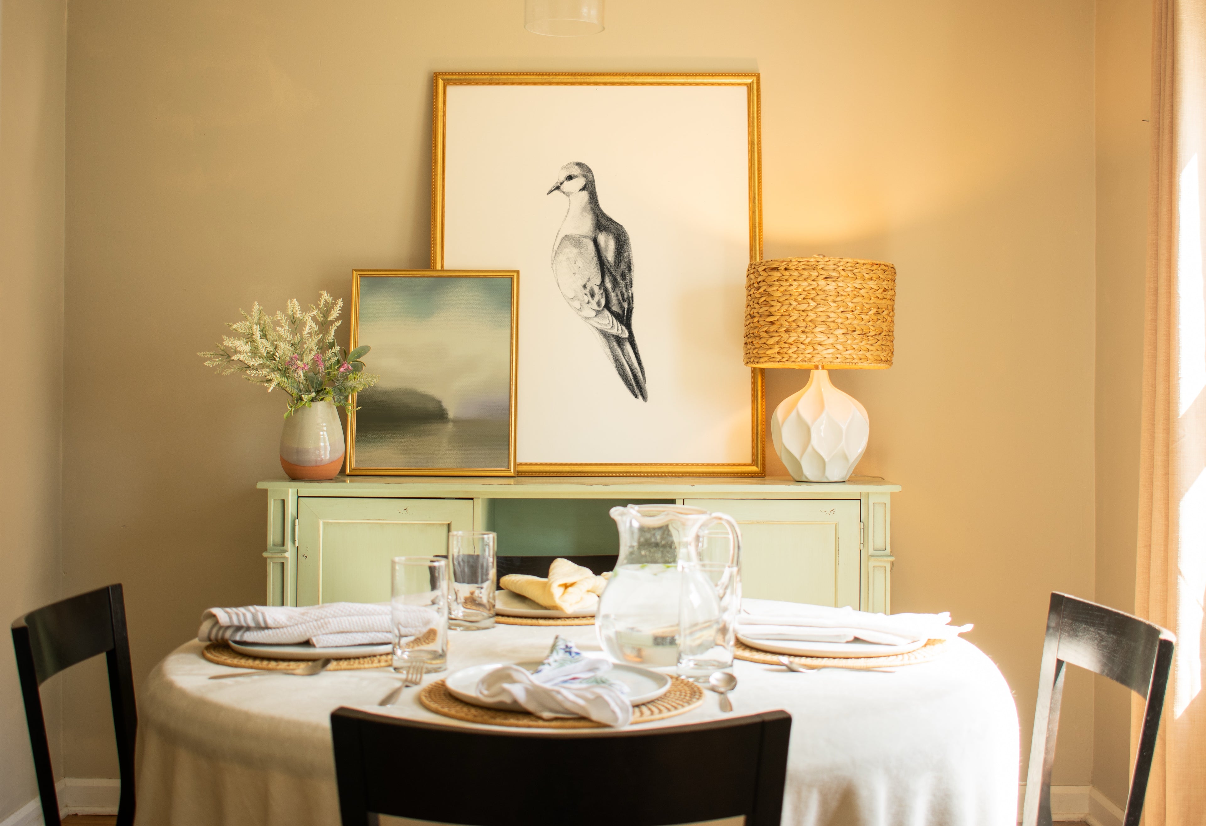 Dining room with a table set for four, framed artwork on the wall, and a lamp.