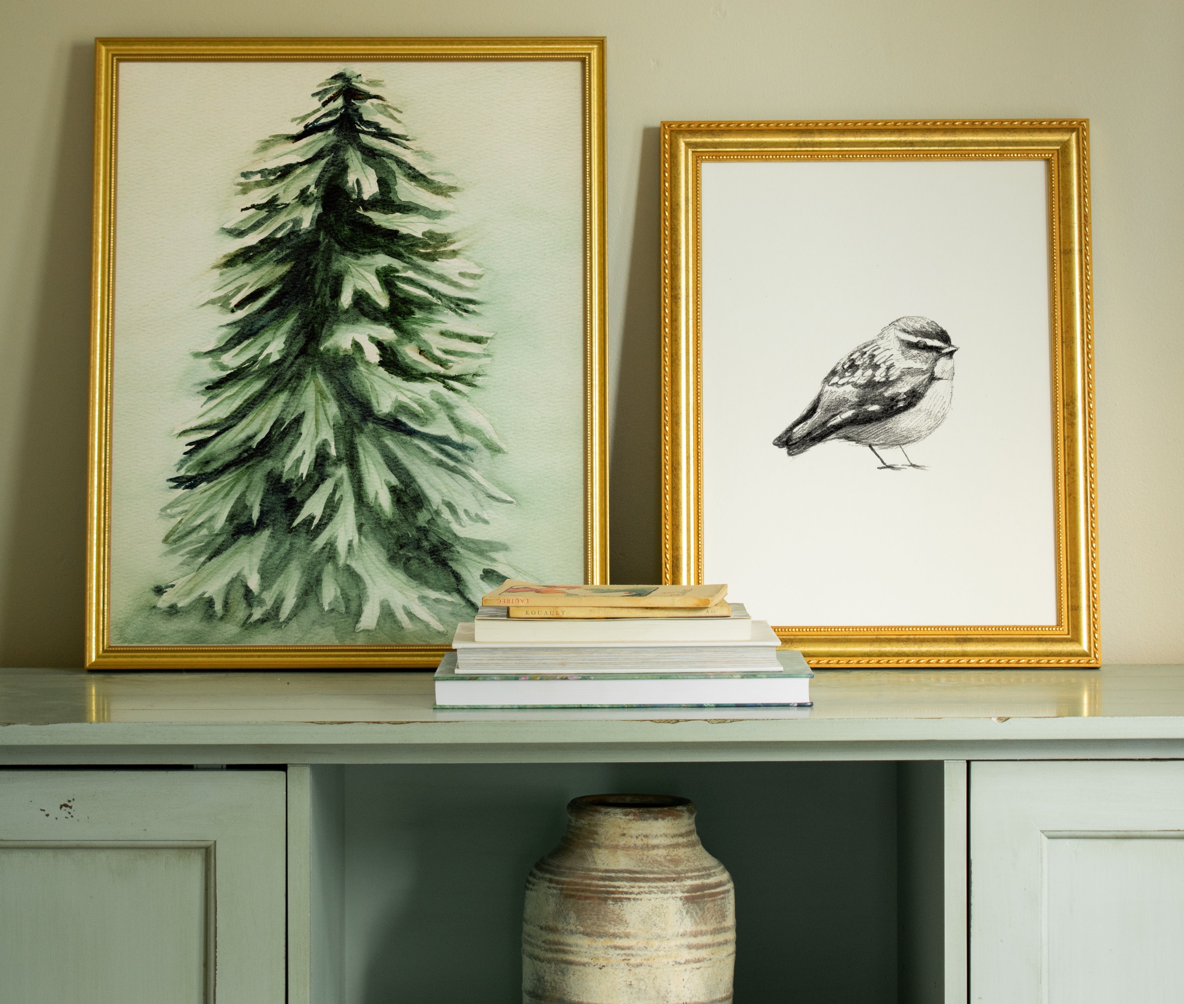 Two framed artworks on a shelf with books and a vase below. Christmas themed decor.
