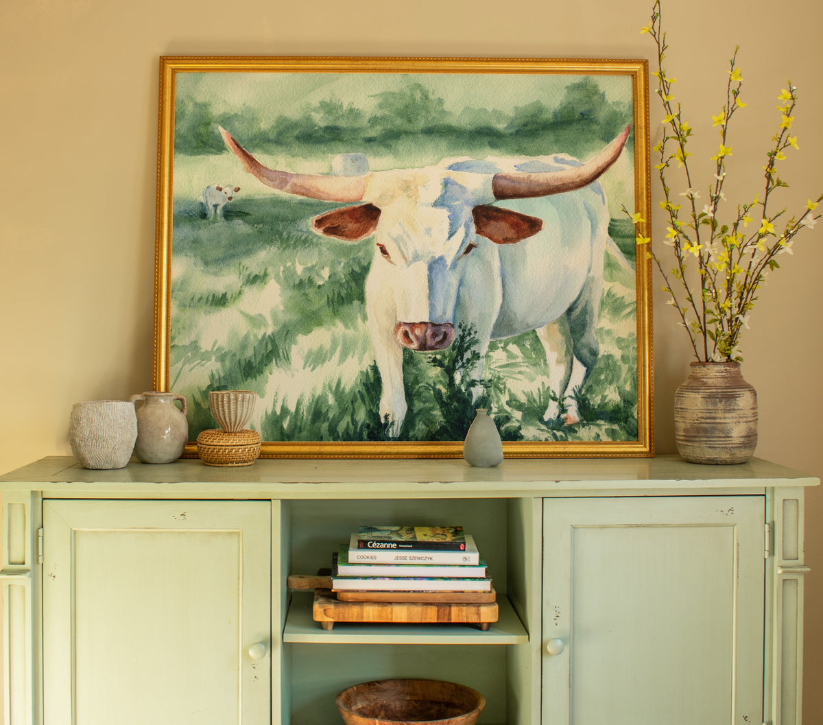 A framed painting of a white longhorn cow in a green field, displayed on a pale sage green wooden sideboard against a beige wall.