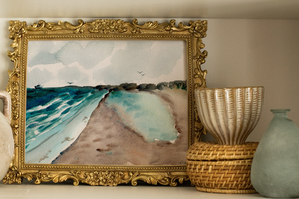 A framed watercolor painting of a beach and ocean waves, set in an ornate gold frame and displayed on a white shelf alongside decorative ceramic vases and baskets.